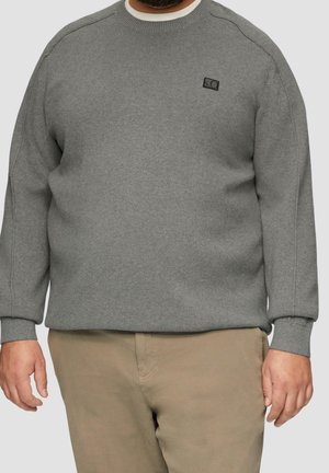 Mand iført en ensfarvet grå crewneck-trøje med en lille sort logo på brystet og beige bukser mod en grå baggrund.