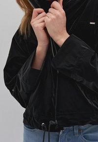 Person som bär en svart zip-up-jacka och blå jeans, som håller i jackans krage med båda händerna nära ansiktet.