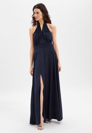 Femme aux cheveux ondulés portant une robe sans manches bleu marine à col licou avec une fente haute sur la cuisse et des sandales argentées à talons hauts, debout devant un fond blanc.