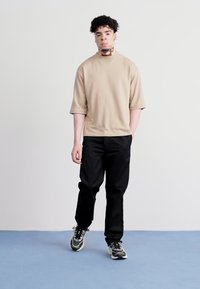 Beige kortærmet sweatshirt med høj hals, parret med sorte bukser. Modellen har grå og sorte træningssko på.