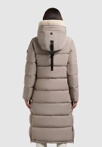 Palton puffer lung de culoare bej, cu glugă căptușită cu blană, model cusut vertical, curea neagră ajustabilă pe spate și buzunare laterale cu fermoar.