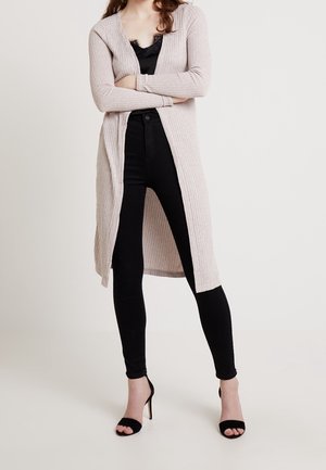 Frau steht mit verschränkten Armen, trägt eine beige lange Strickjacke, ein schwarzes Spitzenoberteil, schwarze enge Hosen und schwarze Sandalen mit hohen Absätzen.