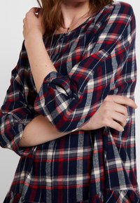 Femme portant une chemise à manches longues à carreaux rouge, bleu marine et blanc, manches retroussées, tenant un bras croisé sur sa poitrine.