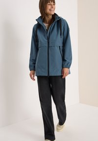 Veste imperméable bleue avec fermeture éclair à l'avant, capuche ajustable, poignets élastiques et coupe décontractée, portée par-dessus un pantalon noir et des baskets légères.