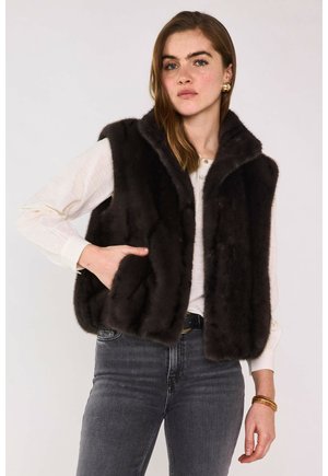 Donna che indossa un gilet di pelliccia nero sopra una camicia bianca a maniche lunghe e jeans grigi, in piedi con una mano nella tasca del gilet contro uno sfondo semplice.