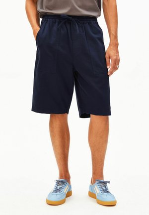 Homme portant un short à cordon bleu marine, une chemise grise rentrée, et des baskets bleu clair avec des semelles en gomme, debout les mains partiellement dans les poches.