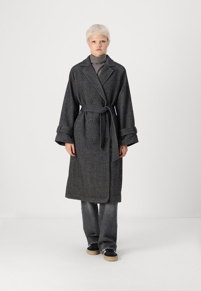 Trench coat de espiga gris con diseño envolvente, cintura ceñida con cinturón, grandes solapas y puños anchos. Combinado con un jersey de cuello alto gris y vaqueros de campana.