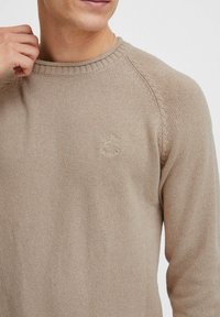 Lysbrun strikket sweater med rund hals, raglanærmer og en lille logo detalje på brystet. Blød tekstur med en fin vævning.