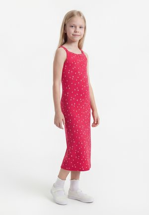 Mergina stovinti dėvi raudoną suknelę be rankovių su mažais baltais gėlių raštais, baltas kojines ir baltus batus ant paprasto balto fono.