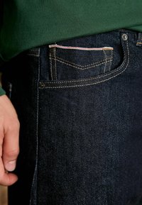 Jeans din denim închise la culoare, cu estompare subtilă, design cu cinci buzunare, cusături aurii și un mic accent roșu pe buzunar. Poziționate aproape de un top verde.