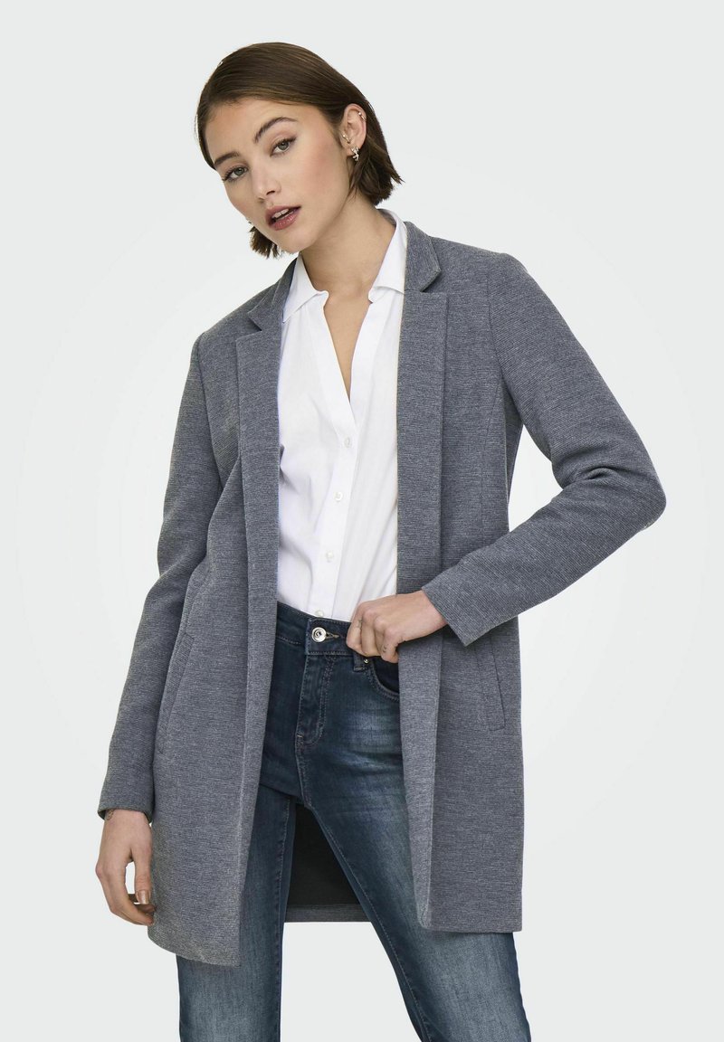 Cardigan long gris tissé avec une ouverture à l'avant, associé à une chemise blanche à col et à un jean en denim. Présente une coupe décontractée et un design minimaliste.