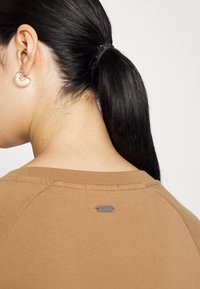 Brown cotton top with a crew neckline and shoulder seams. A small branded tag is visible at the back near the neckline. Smooth texture.