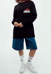 Svart långärmad t-shirt med färggrafik "GRIMY", som kombineras med blå denimshorts och vita sneakers med mönstrade strumpor.