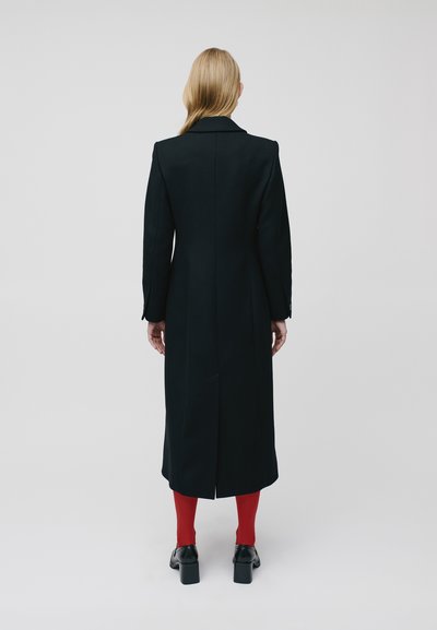Palton lung negru cu guler ascuțit, croială dreaptă și fante laterale. Purtați cu ciorapi roșii și pantofi cu toc negri. Design minimalis.