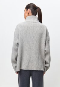 Helle graue Sweater mit vertikaler Rippstruktur, hohem Kragen, überschnittenen Schultern und einem leicht oversized Schnitt. Kombiniert mit dunkelgrauen Hosen.
