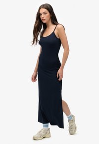 Robe maxi sans manches bleu marine avec une silhouette ajustée et une fente sur le côté, réalisée dans un matériau doux. Assortie à des baskets beiges et des chaussettes bleu clair.