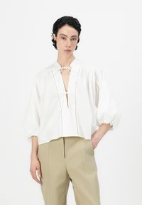 Blusa bianca cropped con maniche a sbuffo, chiusura con fiocco al collo e pattina verticale. Tessuto morbido, design semplice con dettagli di cucitura sottili.