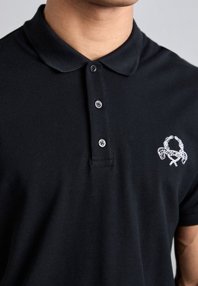 Svart polo med strukturert tyg, treknappslukning og vit brodert logo som har laurbærblad og krossede gjenstander.