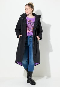 Svart lång kappa med huva, kombinerad med en lila grafisk sweatshirts och blå jeans. Glansiga svarta kängor fulländar outfiten.