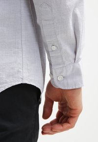 Gros plan sur le bras droit d'une personne portant une chemise gris clair à manches longues avec des poignets boutonnés, suspendu à côté d'un pantalon noir.