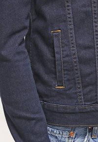 Granatowa kurtka dżinsowa o wysmuklonym fasonie, widocznych szwach, bocznej kieszeni i teksturowanej tkaninie. Kończy się na wysokości pasa.
