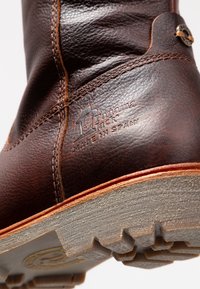 Bottines en cuir marron avec une surface texturée, des coutures contrastantes en beige, et une semelle grise robuste avec un motif de crampons ; marqué "Fabriqué en Espagne."