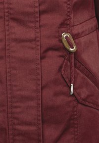 Veste bordeaux en tissu robuste présentant une surface texturée, des poches latérales, un cordon de serrage avec des accessoires dorés, et des coutures renforcées.