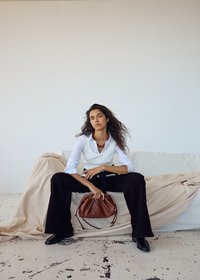 Femme aux longs cheveux bouclés assise sur un canapé couvert, vêtue d'une chemise blanche, d'un pantalon noir et tenant un sac à main marron devant elle.