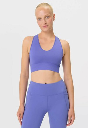 Femme aux cheveux blonds portant un soutien-gorge de sport violet assorti et un legging taille haute, debout devant un fond clair uni.