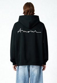 Sudadera negra con capucha que presenta "Amour" bordado en blanco cursivo en la parte posterior. Corte relajado con puños y dobladillo acanalados. Jeans de color azul claro.