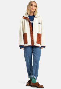 Crème fleece jacket met bruin stoffen accenten, voorzakken en een logo patch. Draagt over een blauwe shirt en gecombineerd met opgerolde denim en bruine schoenen.