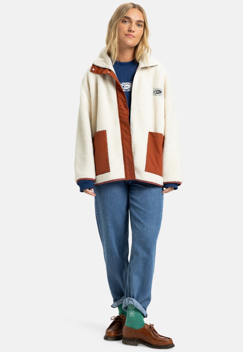 Crème fleece jacket met bruin stoffen accenten, voorzakken en een logo patch. Draagt over een blauwe shirt en gecombineerd met opgerolde denim en bruine schoenen.
