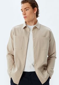 Camisa beige de botones con una tela texturizada, que presenta dos bolsillos frontales y un cuello camisero. Llevada sobre una camiseta blanca de cuello redondo.