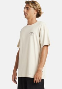 Beige katoenen t-shirt met korte mouwen, ronde hals en geborduurd logo detail aan de voorkant. Ontspannen pasvorm en gladde textuur.