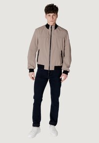 Giacca bomber color beige chiaro con colletto alto, tasche laterali con zip, polsini e orlo inferiore a coste neri. Abbinata a jeans scuri e sneakers bianche.