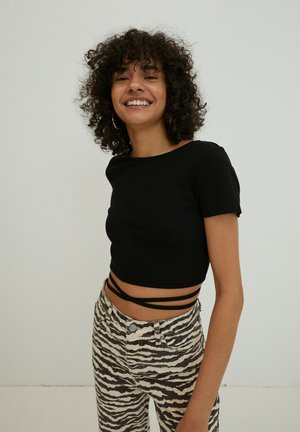 Black ribbed crop top with short sleeves and side straps paired with white and brown striped high-waisted jeans. Plain background.