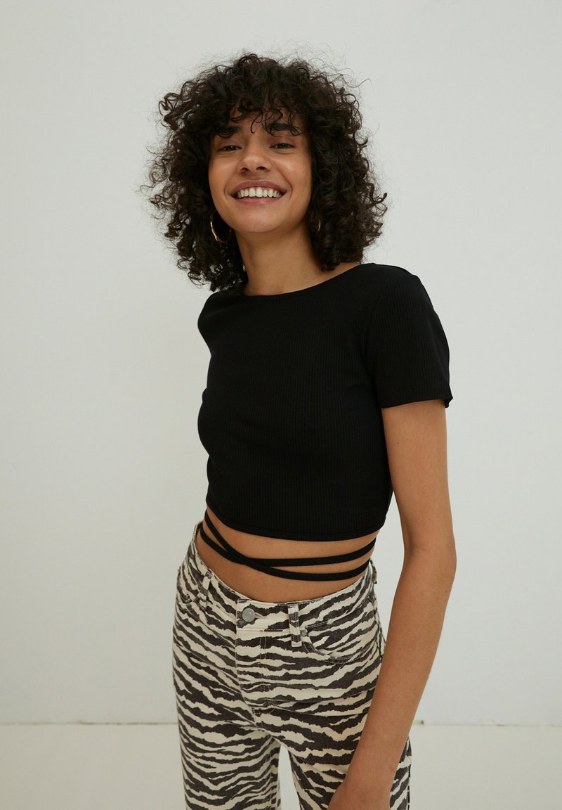 Schwarzes geripptes Cropped-Top mit kurzen Ärmeln und seitlichen Trägern, kombiniert mit hochgeschnittenen Jeans in Weiß und Braun mit Streifen. Schlicher Hintergrund.