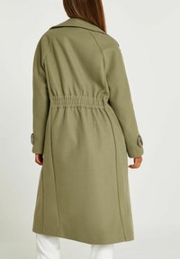 Cappotto lungo verde oliva con vita segnala da cintura, spalle strutturate e polsini abbottonati; realizzato in un tessuto morbido e testurizzato.