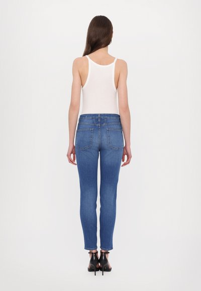 Blå høytlivsjeans med en svak falming, fem lommer og rå kant. Kombinert med en hvit tanktop og svarte hælsandaler.