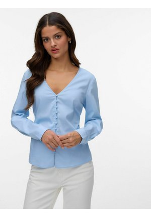 Femme aux longs cheveux bruns portant un chemisier bleu clair boutonné et un pantalon blanc, debout devant un fond blanc uni.