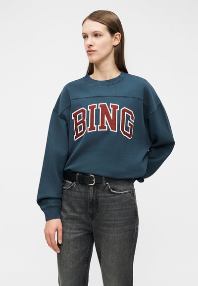 Kvinne med rett brunt hår har på seg en teal genser med teksten "BING", stukket inn i falmede svarte jeans med et svart belte, hendene avslappet.