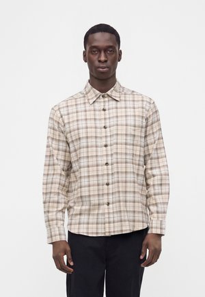 Homme portant une chemise à carreaux beige et marron à manches longues et un pantalon noir, debout devant un fond blanc uni.