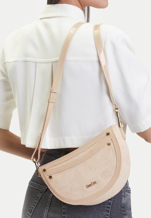 Frau trägt weiße kurze Jacke und schwarze Jeans mit beige-farbener, mondförmiger Wildleder-Umhängetasche mit verstellbarem Riemen.