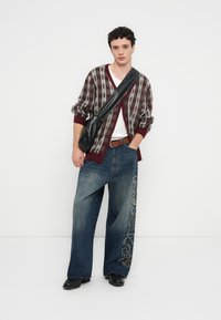 Pleteni kardigan u bordo i sivim prugama preko bijele košulje; široke plave traperice s bogatom vezenom dekoracijom; crna torbica i cipele.