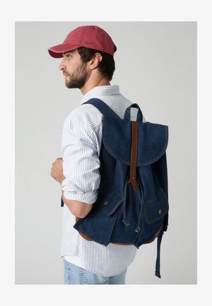 Denim backpack with a flap closure, brown leather accents, blue fabric, and dual front pockets. Adjustable straps, sturdy construction.