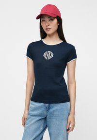 Μπλέ σκούρο εφαρμοστό t-shirt με κοντά μανίκια, λευκή λεπτομέρεια γύρω από τον λαιμό και ένα λογότυπο στο κέντρο. Συνδυάζεται με ανοιχτό μπλε τζιν και μία κόκκινη καπέλο.