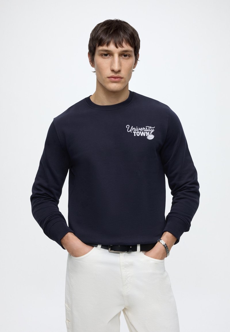 Ung mand med kort mørkt hår iført en sort sweatshirt med teksten "University Town" og hvide bukser, står med hænderne i lommerne.