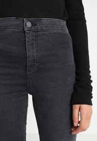 Detailní záběr osoby, která má na sobě vysoce posazené vybledlé černé džíny a dlouhý černý top, přičemž pravá ruka leží vedle těla.