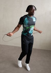 Black athletic shirt with green and blue abstract pattern, paired with black pants and white sneakers; model is skipping with a rope.
