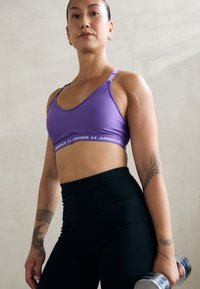 Sujetador deportivo morado con tirantes ajustables, con una banda de logo blanca. Leggings negros de cintura alta. Persona sostiene una mancuerna. Apariencia atlética y suave.